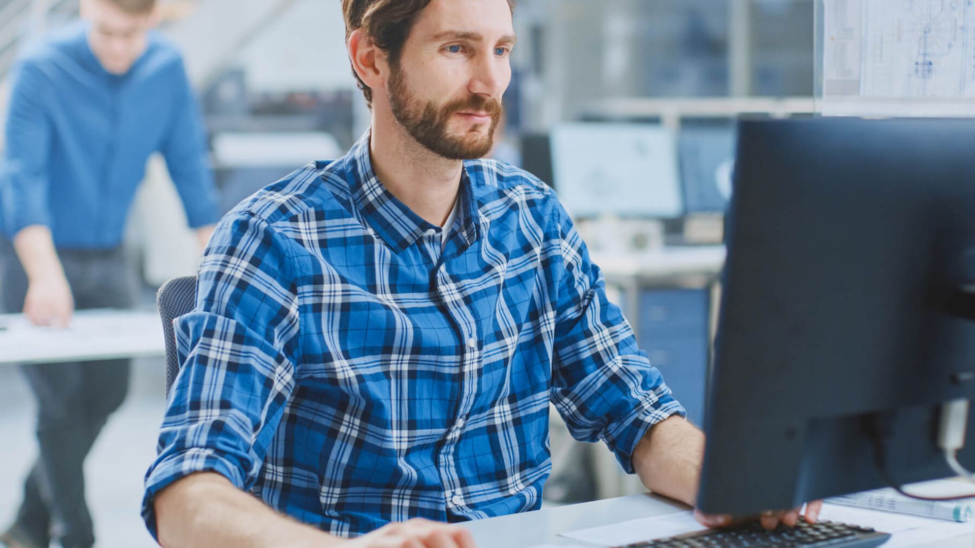 Ein Mann sitz an einem Schreibtisch vor einem Bildschirm und tippt auf der Tastatur. Im Hintergrund ist ein Büro zusehen.