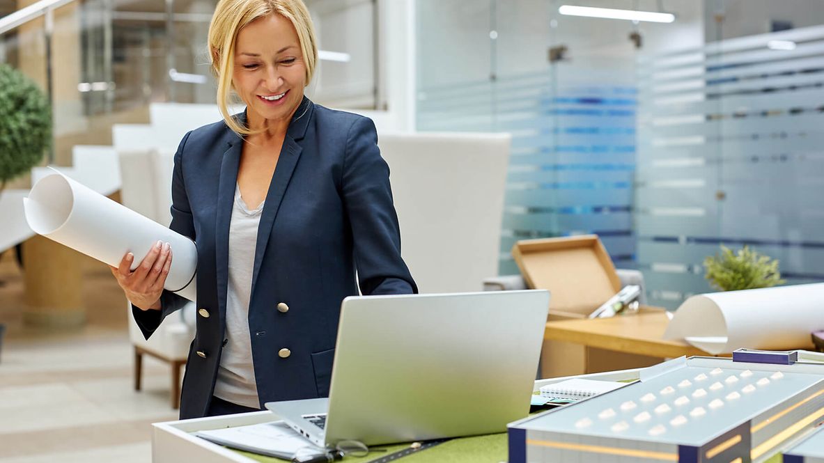 Eine Frau steht in ihrem Büro, unter dem Arm trägt sie eine Papierrolle. 