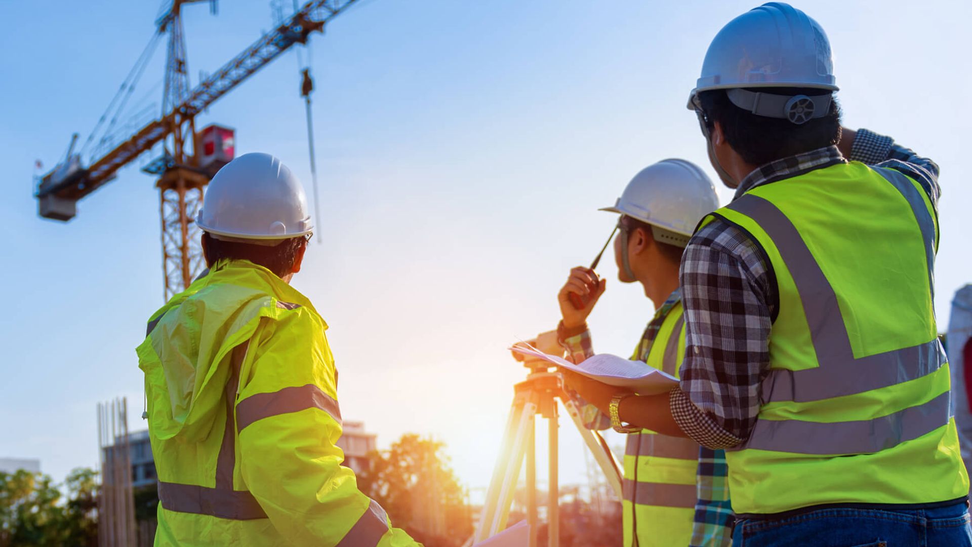 3 Personen mit Warnwesten stehen um ein Vermessungsgerät vor einem Kran.
