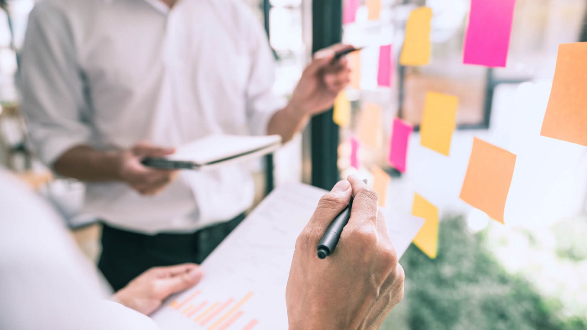 Zwei Personen stehen vor einer Wand mit Pinken und orangenen Post its. Sie halten Notizblätter und Stifte in der Hand.
