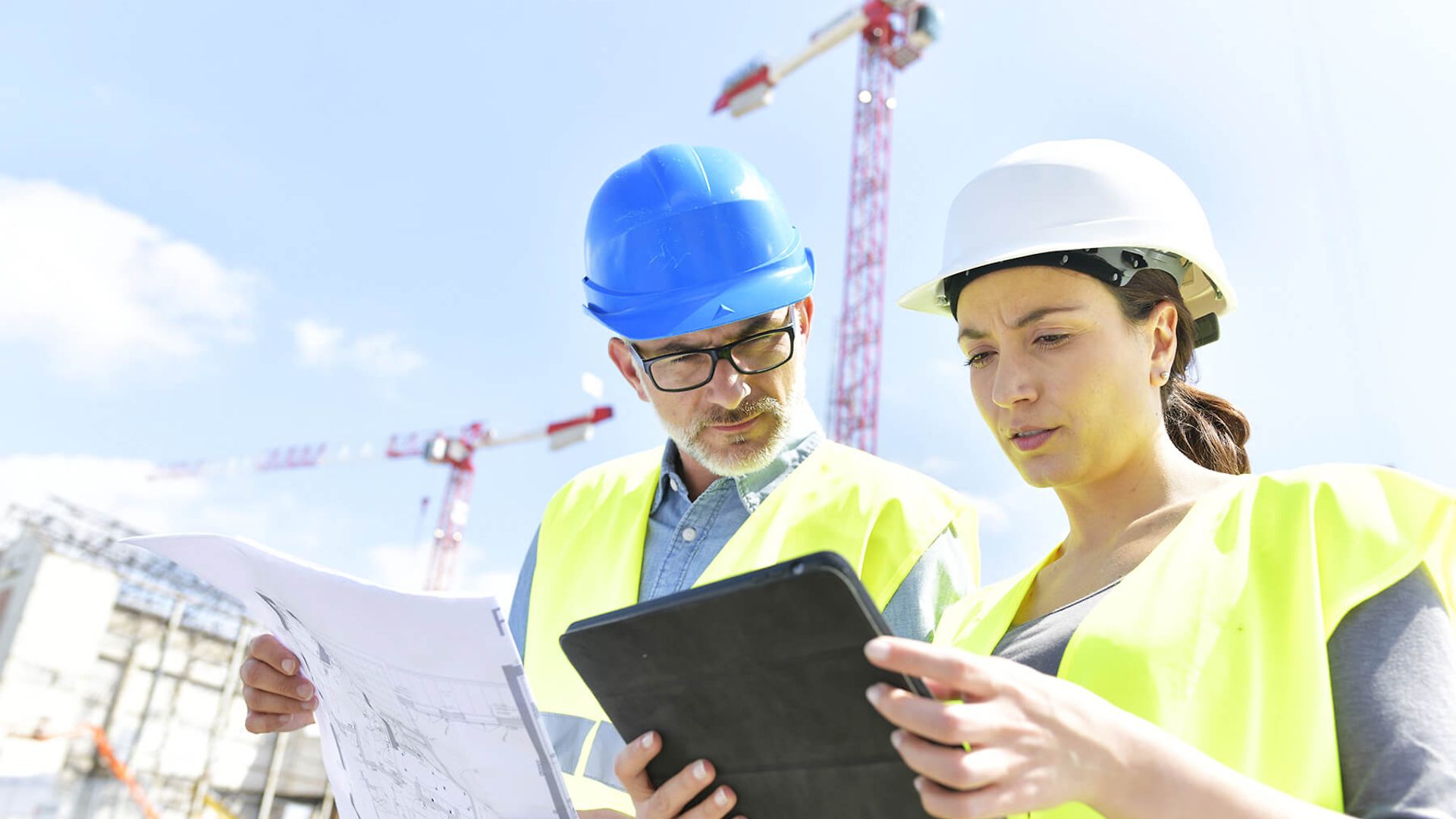 Ein Mann und eine Frau betrachten gemeinsam ein Tablet. Beide tragen Schutzwesten und Helme.