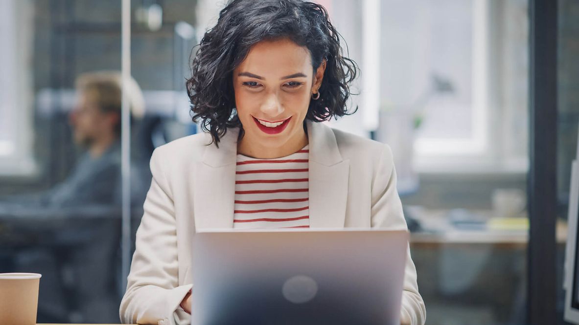 Eine Frau sitzt vor ihrem Laptop und lächelt. Im Hintergrund ist ein Büro zu sehen.