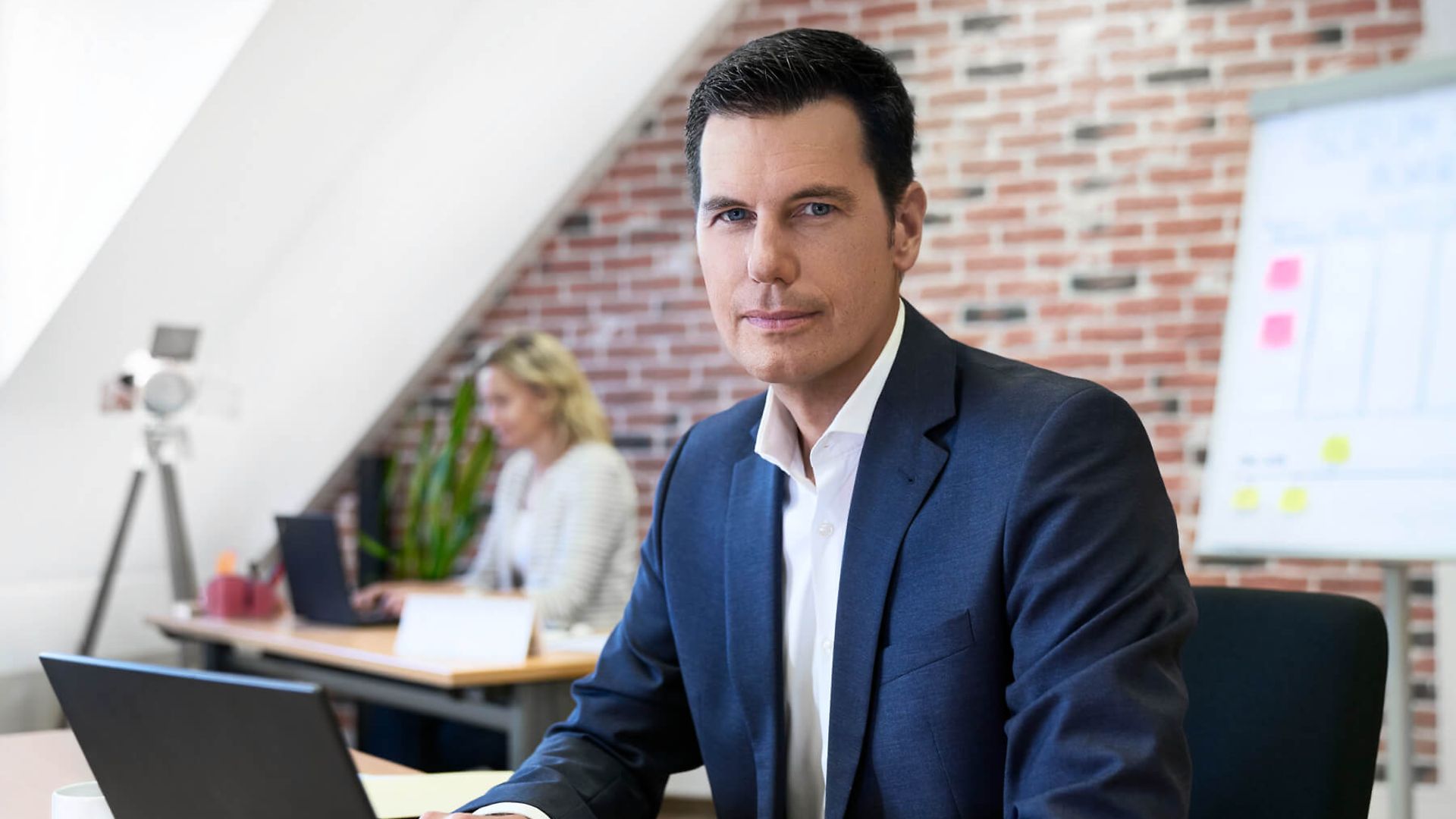 Ein Mann sitzt auf einem Schreibtischstuhl und blickt in die Kamera. Vor ihm steht ein Laptop. 