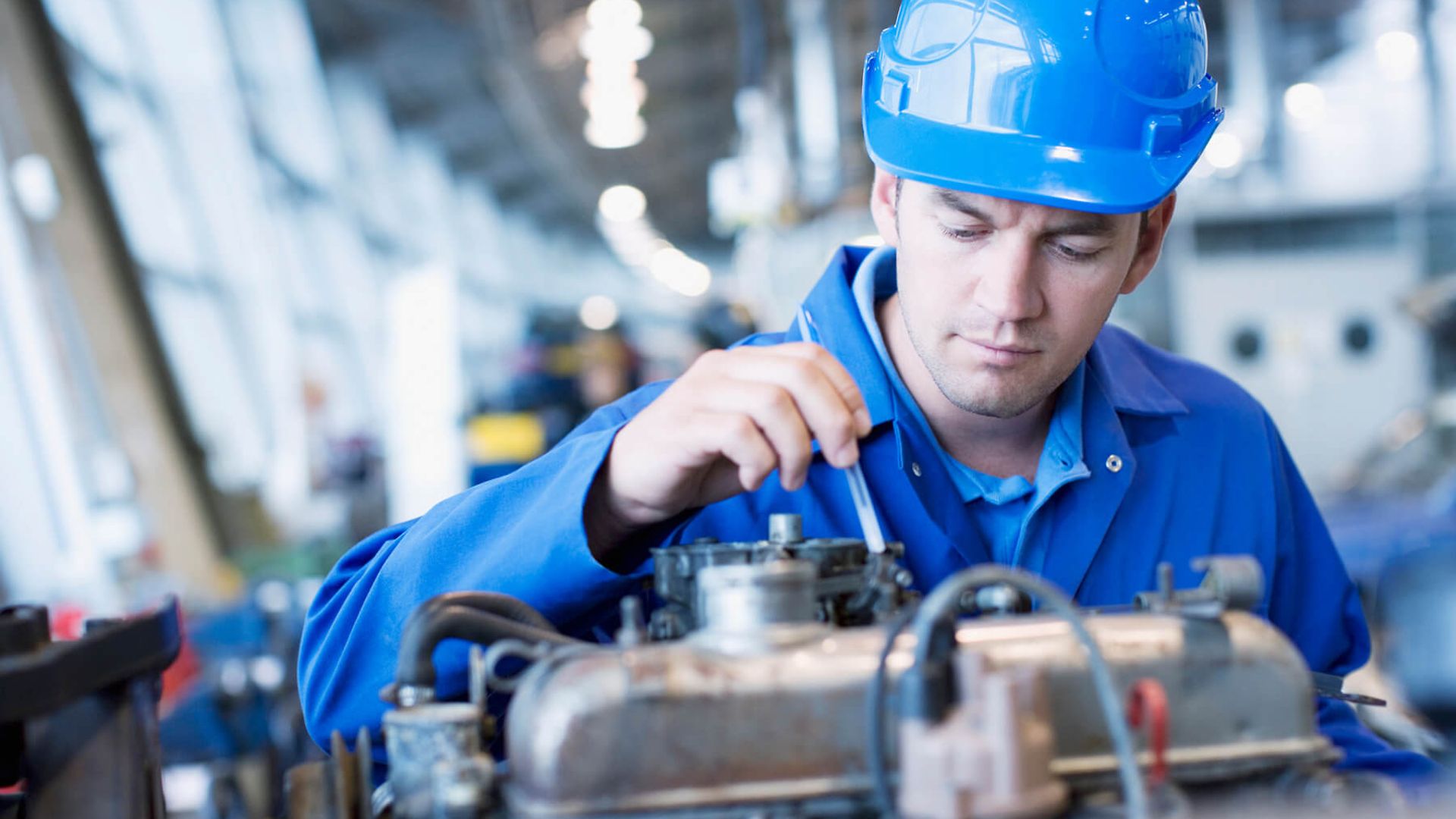 Ein Mann in Blaumann und Helm blickt kritisch in eine Art Tank oder Ölwanne und hält ein Messstäbchen hinein.