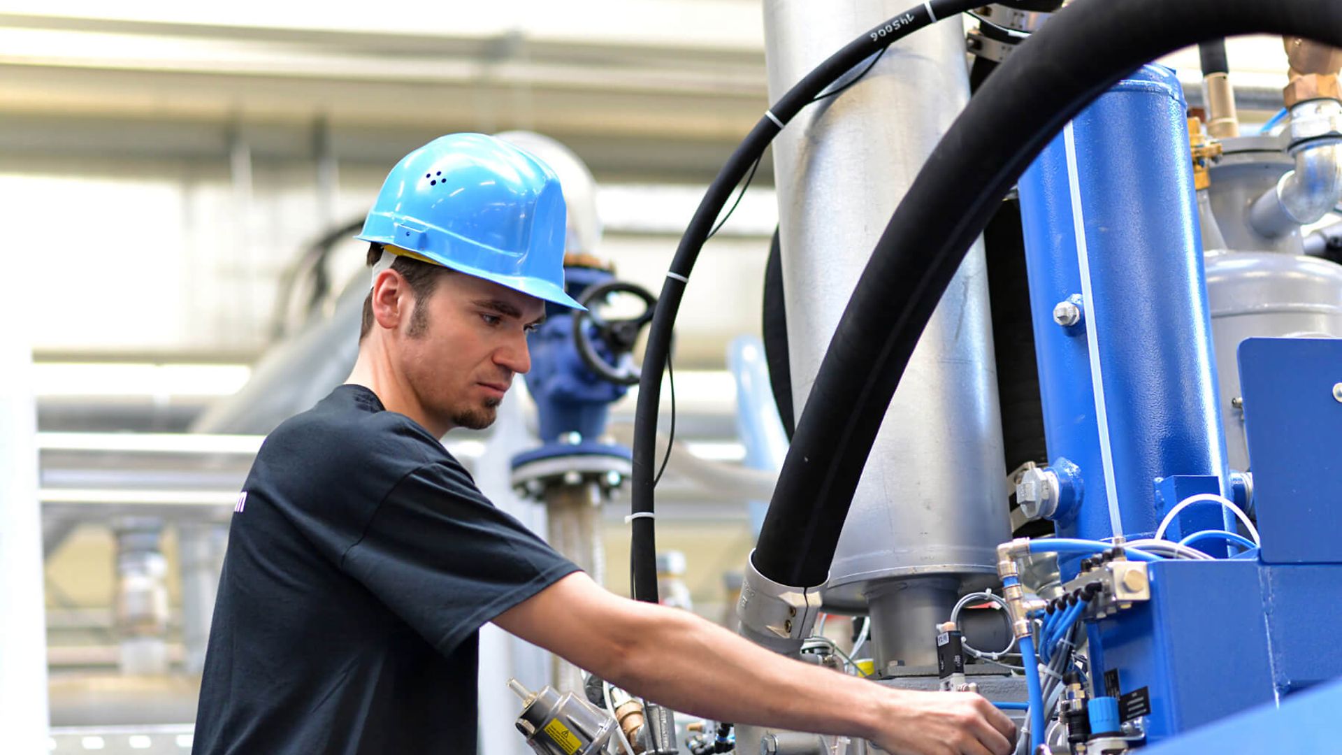 Eine Person mit Schutzhelm bedient eine große Maschine. Im Hintergrund sind Rohre und Ventile zu sehen.