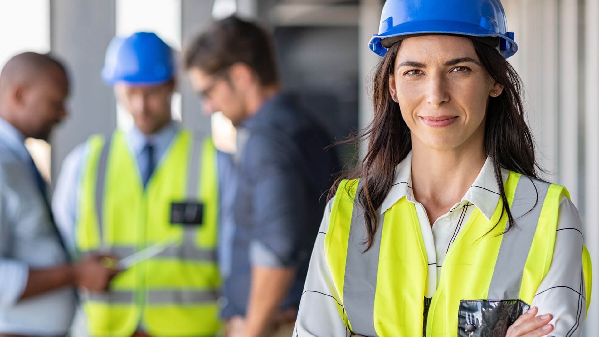 Eine Frau in Arbeitskleidung mit verschränkten Armen steht im Vordergrund und blickt in die Kamera.