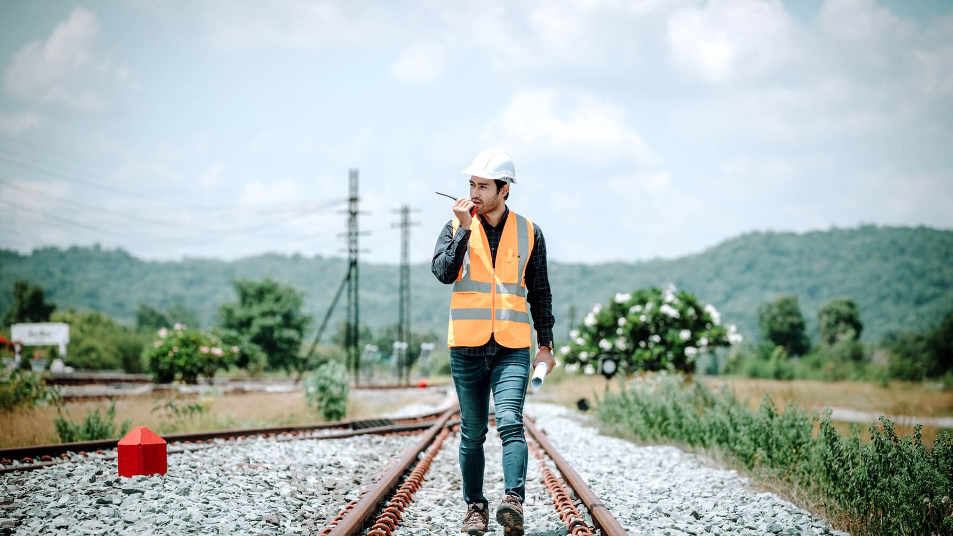 Ein Mann mit Warnweste und Helm läuft zwischen Gleisen in einer ländlichen Umgebung. Er hält ein Funk Gerät an seinen Mund.