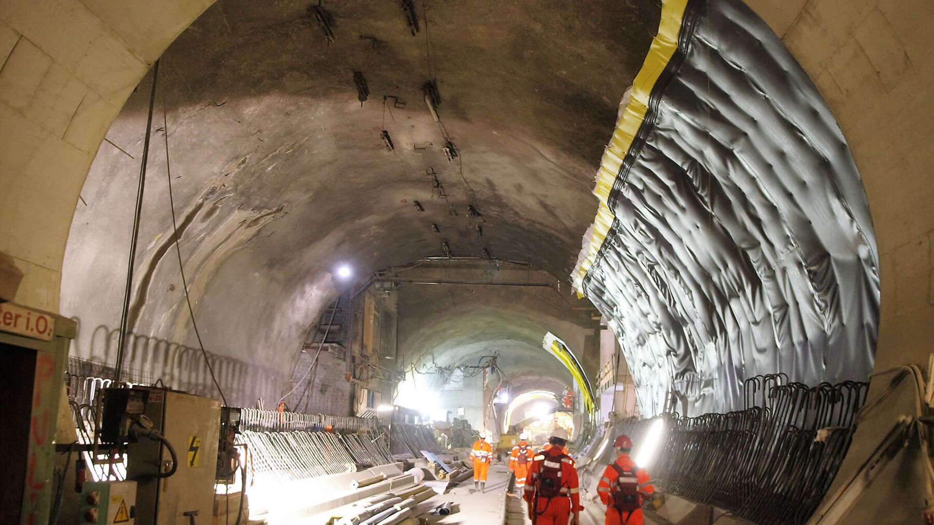 Zwei Personen laufen in einem Rohbau eines großen Tunnels. Beide Personen tragen Schutzkleidung.