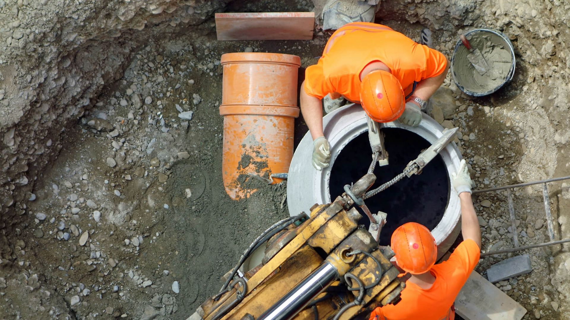Aus der Vogelperspektive sieht man zwei Personen mit orangenen Warnwesten und Helmen, die in einer schlammigen Baugrube stehen