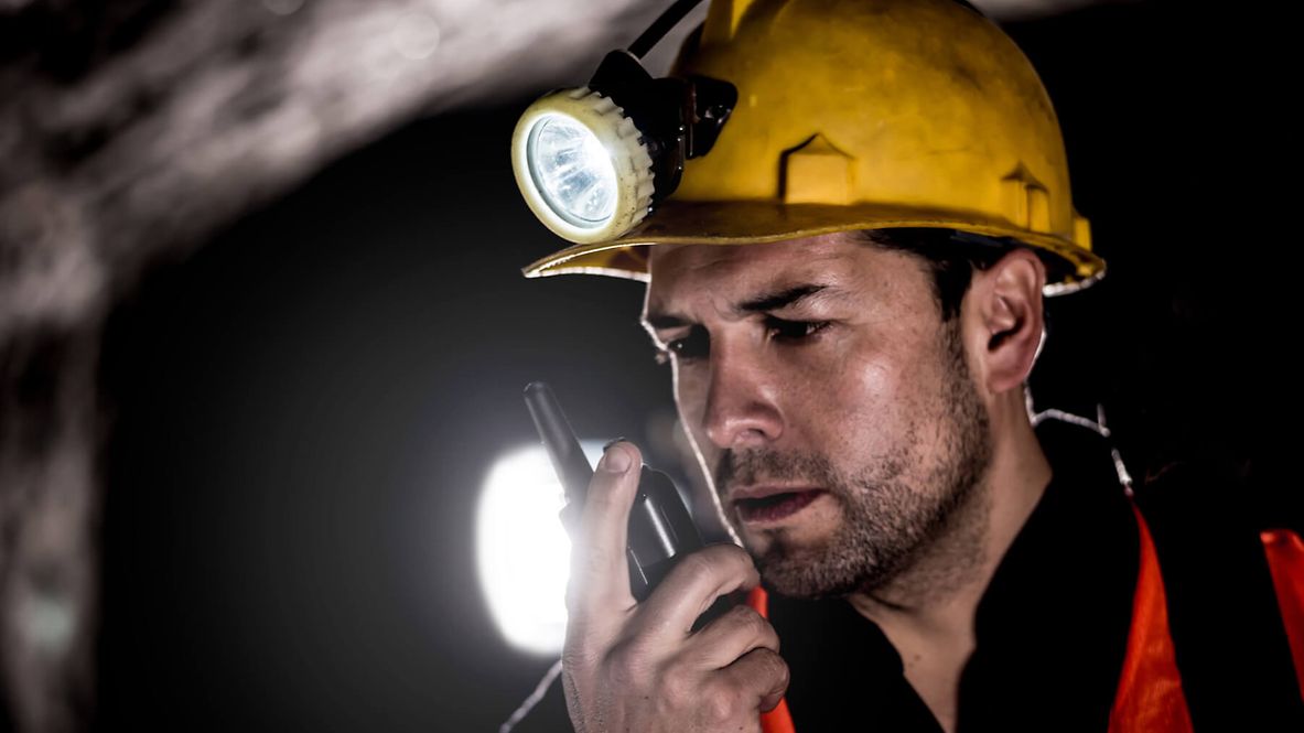Ein Mann der in sein Funkgerät spricht. Er hat eine Lampe auf seinem Helm. Der Hintergrund ist dunkel.