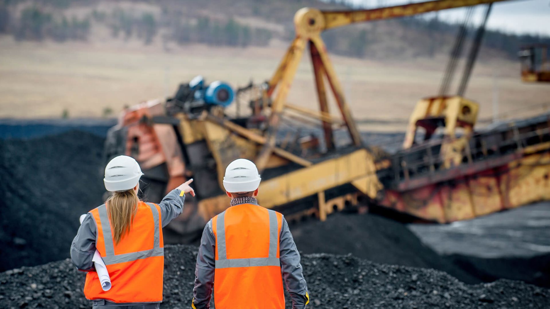 Zwei Mitarbeiter in Warnwesten mit einer Papierrolle unter dem Arm gehen auf einen Schuttberg zu.