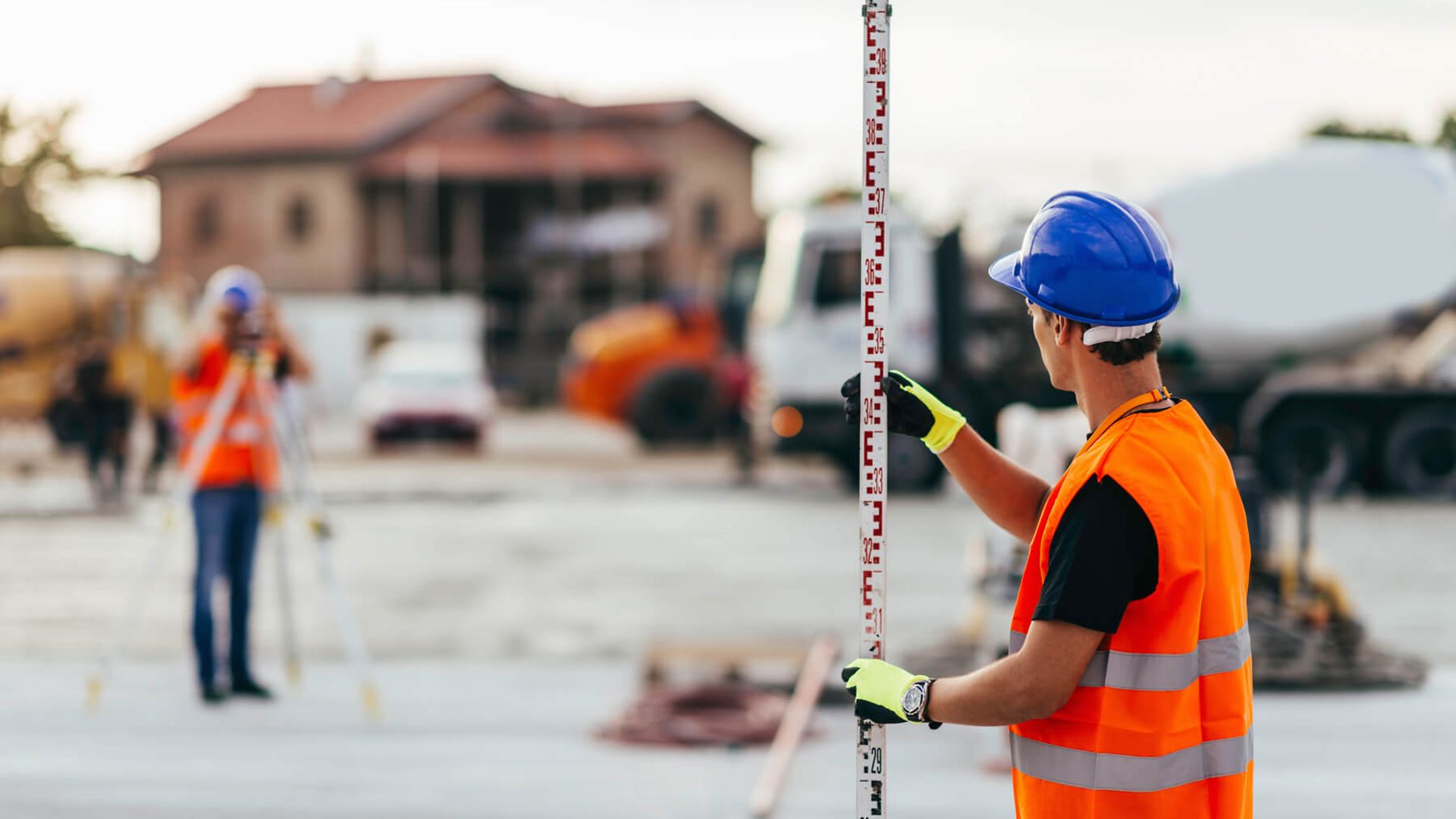 Ein Mann mit Warnweste und Helm steht im Vordergrund. Er hält eine Messlatte senkrecht. 