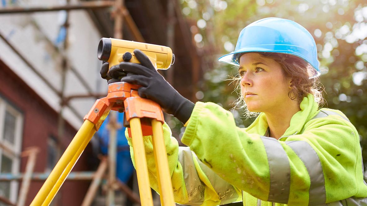 Eine Frau mit Warnweste und Helm blickt durch ein Landvermessungsgerät.
