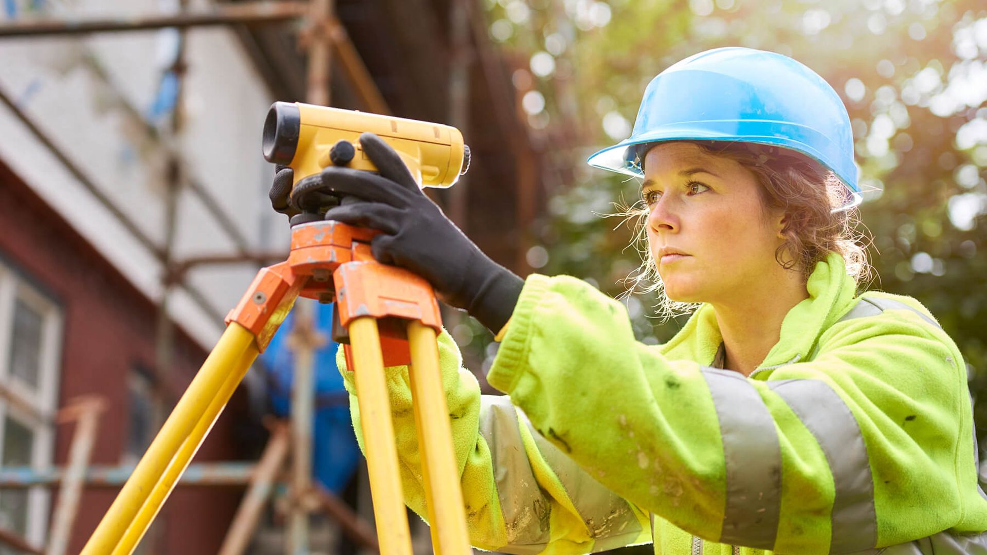 Eine Frau mit Warnweste und Helm blickt durch ein Landvermessungsgerät.