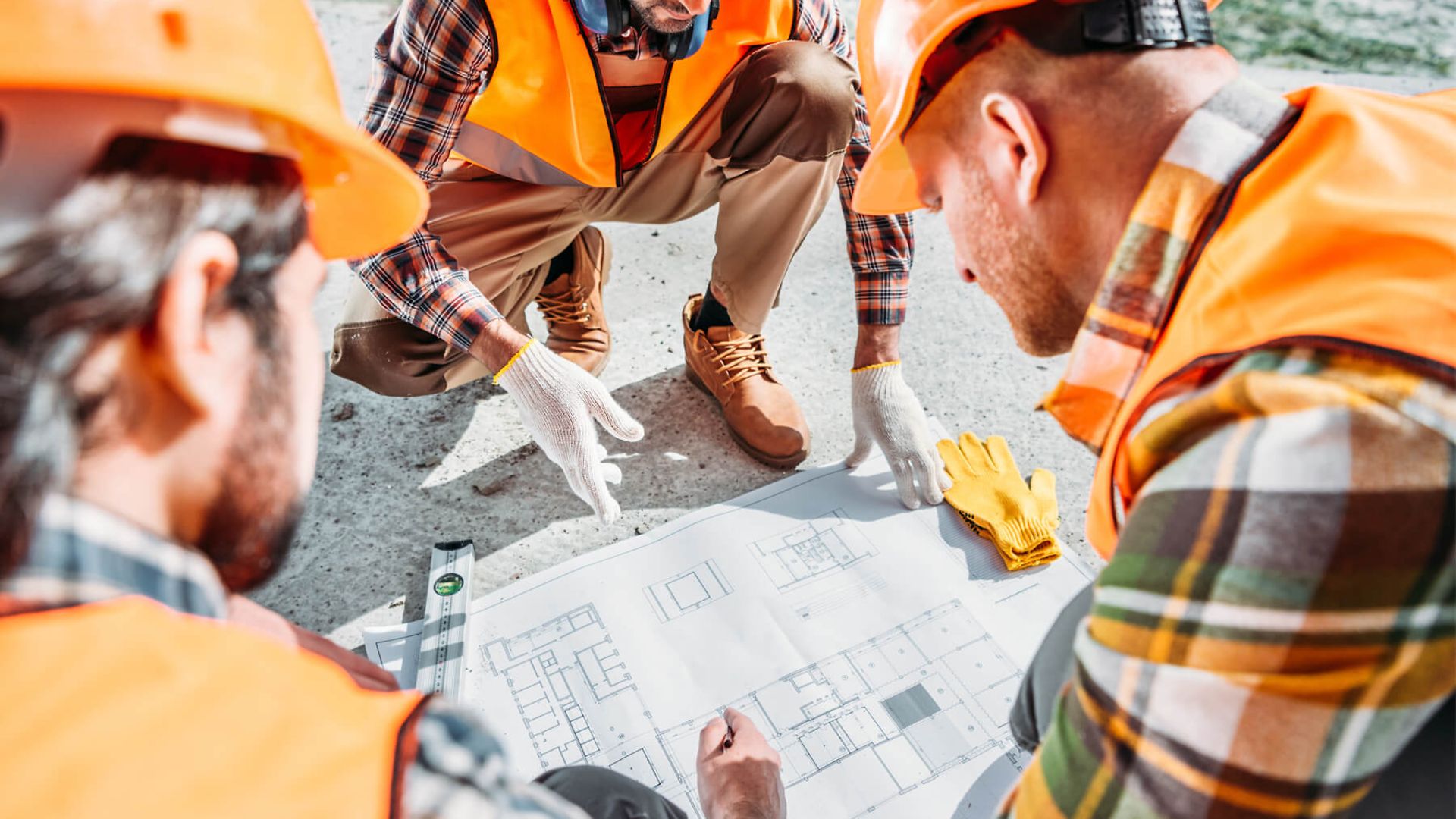 Mehrere Personen in orangenen Warnwesten und Helmen unterhalten sich über den Grundriss, der vor ihnen auf dem Boden liegt.