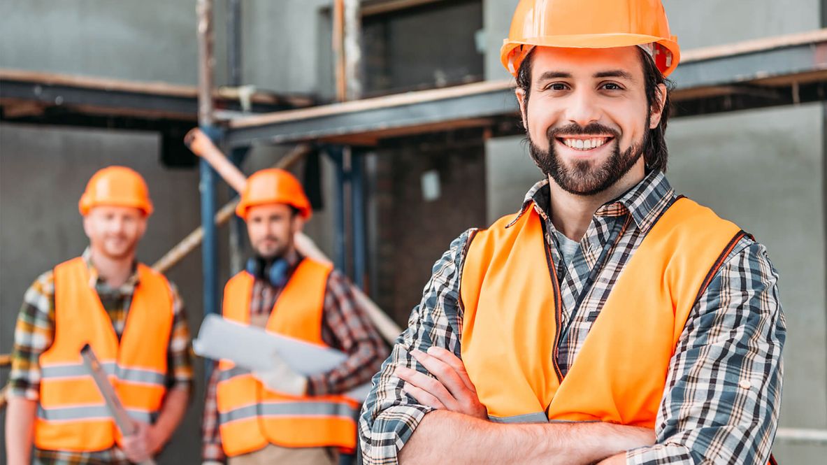 Auf dem Bild sind drei Personen in Schutzkleidung vor einem Gerüst. Der Mann im Vordergrund lächelt in die Kamera.