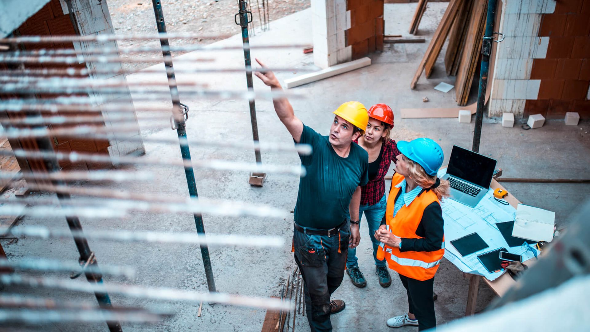 Drei Personen mit Schutzhelmen stehen vor einem provisorischen Arbeitstisch, auf dem ein Laptop steht und Notizen liegen