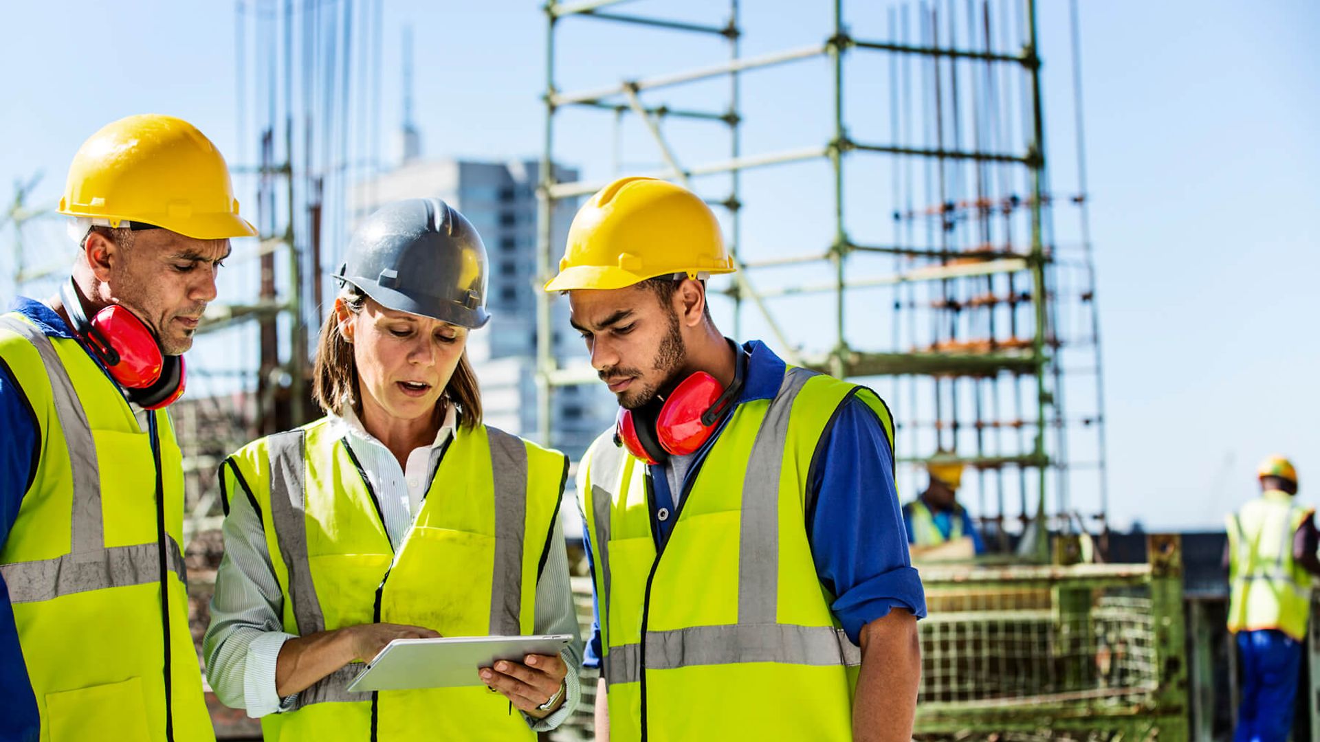 Drei Personen mit Warnweste und Helm beugen sich über einen Notizblock. Im Hintergrund ist eine Baustelle zu sehen.