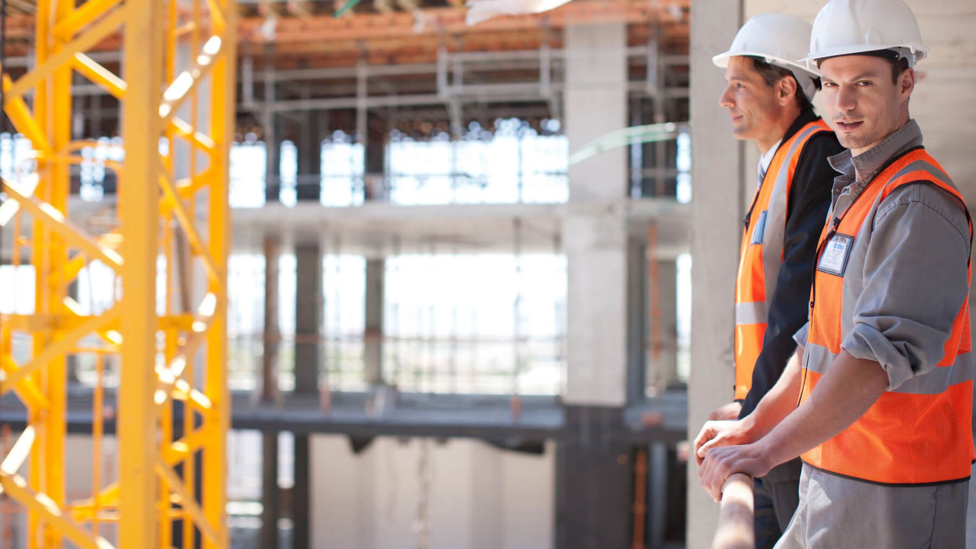 Zwei Personen mit Schutzkleidung stehen in einem Rohbau eines Gebäudes und sehen sich um.