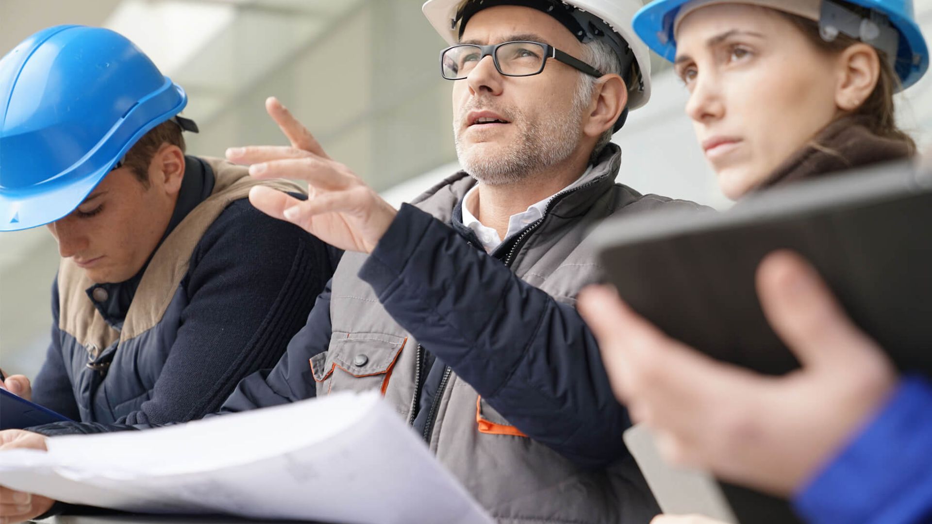 Drei Personen mit Helmen, Plänen und Tablets stehen zusammen und unterhalten sich.