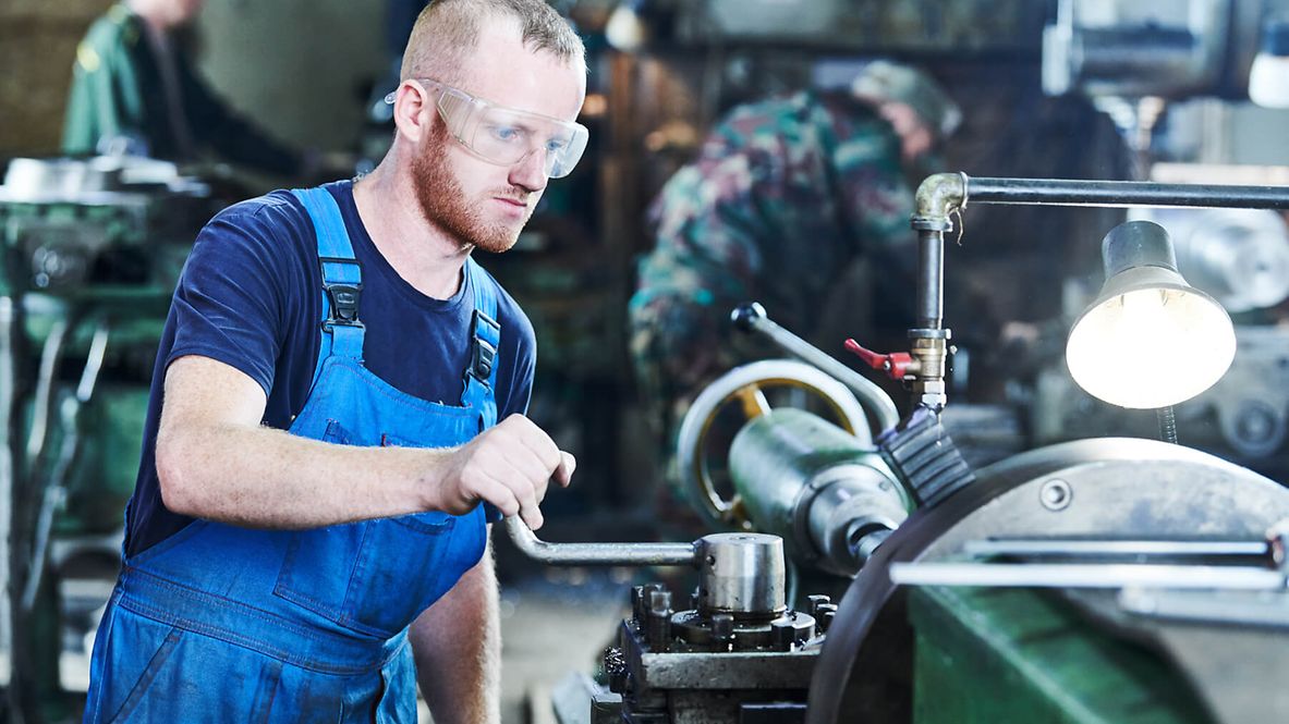 Ein Mann in Arbeitskleidung und Schutzbrille steht an einer großen Maschine und bedient einen Hebel.
